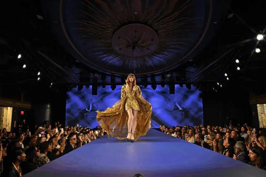 A model in a flowing gold gown on a runway lit in blue before a packed audience